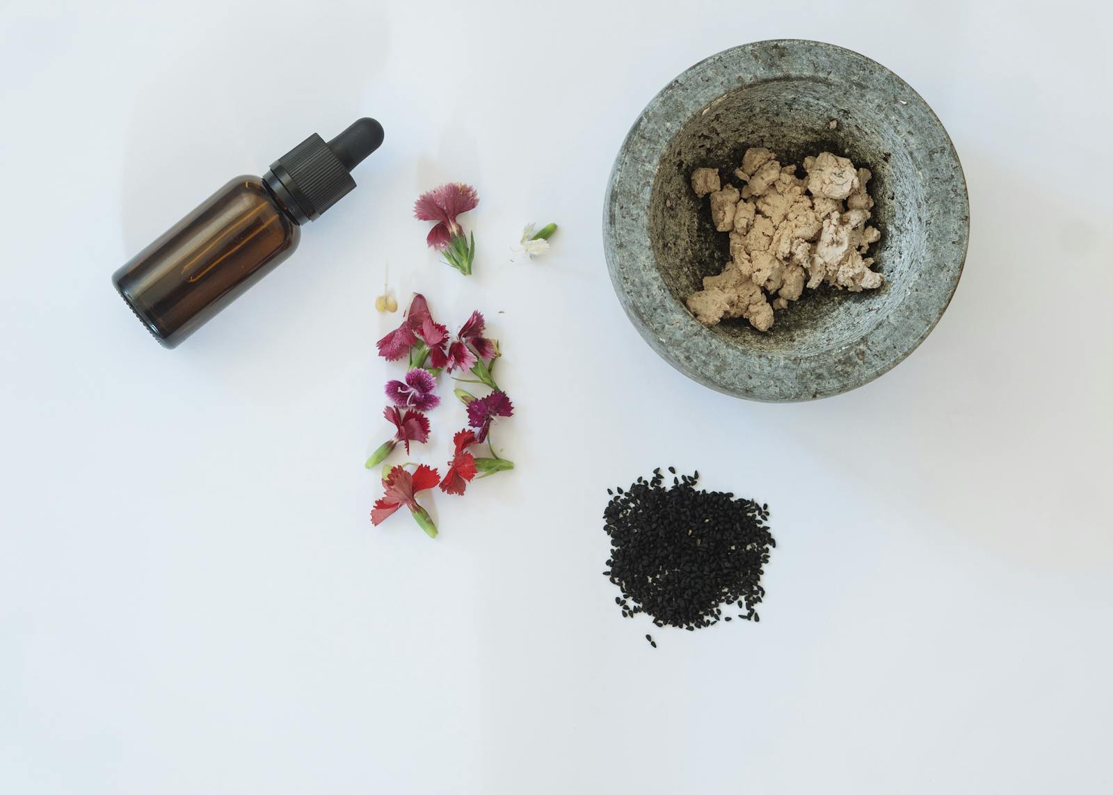 Flat lay of natural skincare ingredients including flowers, black seeds, and a dropper bottle.